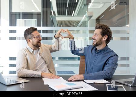 Two smiling men forming a heart shape with their hands, expressing love, collaboration, and friendship while working together in a modern office meeting room Stock Photo