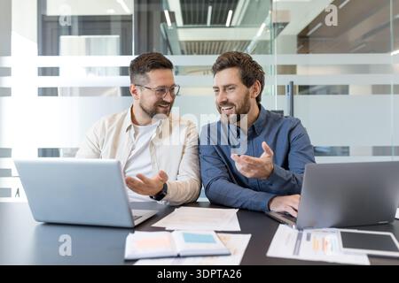 Two smiling male business colleagues are collaborating on a project, actively discussing ideas and sharing insights while working with laptops and documents in a contemporary office environment Stock Photo