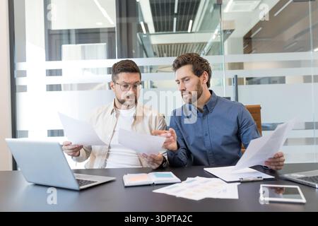 Two male business professionals are reviewing documents and discussing a project, collaborating effectively at a conference table with laptops and papers in a contemporary corporate office setting Stock Photo