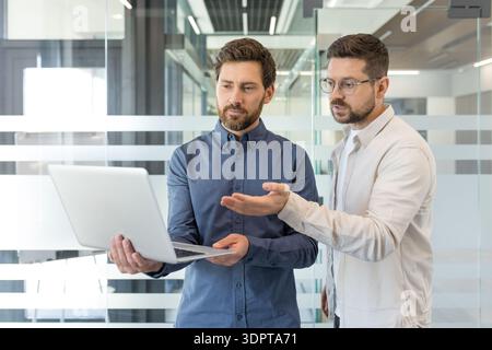 Two business colleagues in a modern office collaborate over a laptop, discussing strategy and ideas, pointing at the screen and planning a project with confident, professional focus Stock Photo