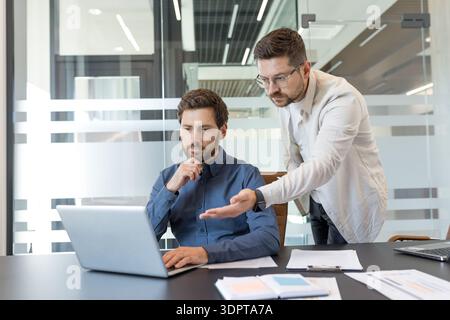 Business colleagues collaborating on a laptop, discussing project details and analyzing data in a bright, contemporary office environment, demonstrating teamwork and professional partnership Stock Photo