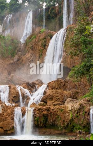 Multiple streams of Thi Lo Su Waterfall cascade over layered limestone rock surrounded by dense tropical forest. Stock Photo