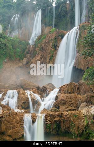 Multi-tier waterfall flowing over rugged rock formations with mist rising through surrounding forest vegetation. Stock Photo