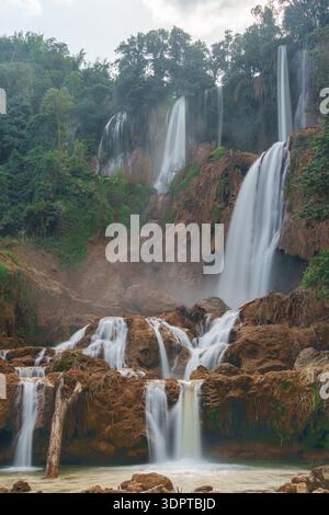Multi tier waterfall flowing over rocky ledges with mist rising among surrounding forest vegetation. Stock Photo