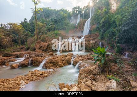 A multi tiered waterfall cascades over rocky formations into shallow river pools surrounded by dense tropical forest. Stock Photo