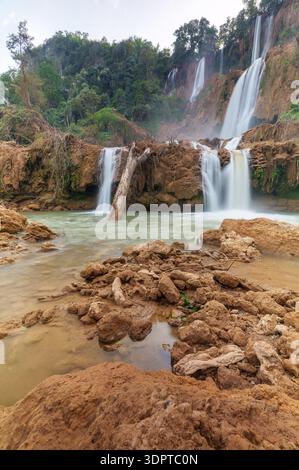 A wide multi tier waterfall cascades over brown rock ledges into a shallow river pool, surrounded by dense tropical forest. Stock Photo