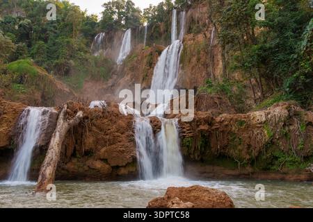 A large multi tier waterfall cascades over brown rock ledges into a fast-moving river, surrounded by dense tropical forest. Stock Photo