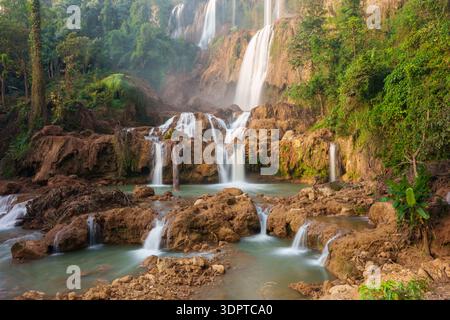 A large multi tier waterfall flows over layered brown rock ledges into multiple shallow pools, surrounded by dense tropical forest. Stock Photo