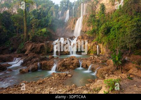 A large multi tier waterfall descends over layered brown rock formations into multiple shallow pools, surrounded by dense tropical forest. Stock Photo