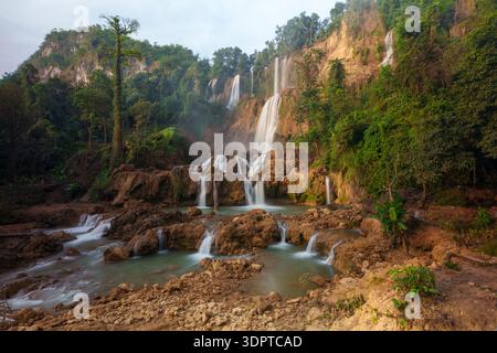 A large multi tier waterfall flows over layered brown rock formations into multiple shallow pools, surrounded by dense tropical forest. Stock Photo