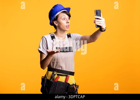 Female construction worker gestures while video calling architects on smartphone. Skilled renovator uses mobile connectivity to remotely receive instructions and coordinate tasks on building job site. Stock Photo