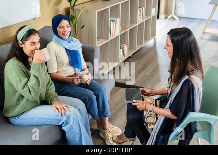 Indian women engaging in discussion on gray sofa in office lounge with coffee mugs and tablet Stock Photo
