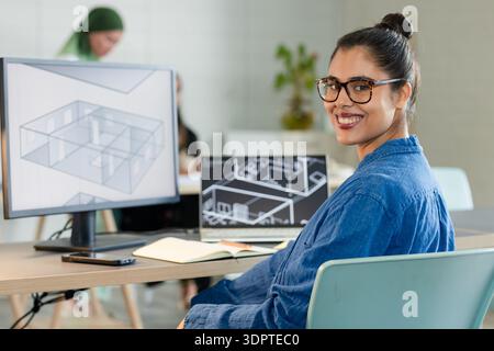 Woman sitting at desk in office working on floor plans on desktop monitor with notebook, smartphone Stock Photo
