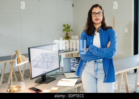 Woman standing arms crossed at worktable in studio reviewing 3D floor plans on monitor and laptop Stock Photo