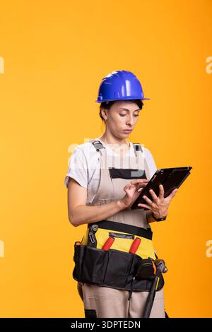 Caucasian female contractor checks digital plans on tablet with focused expression, symbolizing planning or project review. Woman renovator wearing tool belt stands in studio against yellow backdrop. Stock Photo