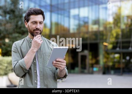 Man looking at a digital tablet and touching his chin while contemplating new ideas or business strategies outside a contemporary office building in the city Stock Photo