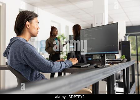 Focused software programmer works in diverse office, ensuring smooth web development. Young adult man types at computer, fully concentrated and committed to completing coding projects efficiently. Stock Photo