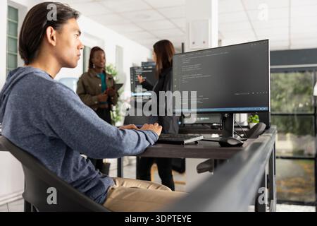 Asian male developer works diligently to improve applications, reviewing backend data on computer. Young adult programmer analyzes HTML coding scripts and focuses on problem solving and optimization. Stock Photo
