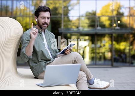 Young man using a laptop and notebook for remote work, participating in an online video conference outdoors while sitting on a modern bench in an urban setting Stock Photo