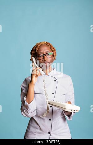 Serious woman chef in uniform listens intently on phone during restaurant business conversation. Female cook with landline receiver, stays focused while receiving directions from supervisor on call. Stock Photo