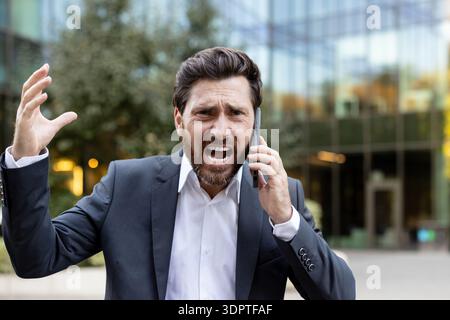 Stressed businessman in a suit having an angry phone conversation outdoors, expressing frustration and anger while using a smartphone in an urban corporate environment Stock Photo