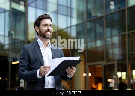 Confident male executive standing in front of a modern glass office building, holding a clipboard with documents and looking thoughtfully into the distance, representing success and future planning Stock Photo