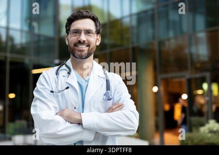 Confident male doctor standing with crossed arms, wearing a white lab coat and stethoscope, smiling while looking at the camera outdoors in front of a modern building Stock Photo