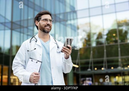 Male doctor in white coat and stethoscope smiling while using a smartphone and holding a clipboard with medical documents, representing modern healthcare professionals Stock Photo