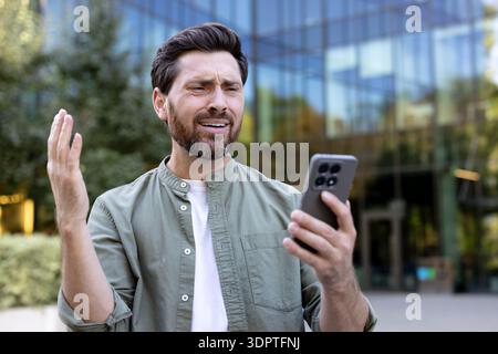 Man experiencing frustration and dissatisfaction while using his smartphone, receiving bad news, looking upset with his hand raised, standing in front of a modern office building Stock Photo