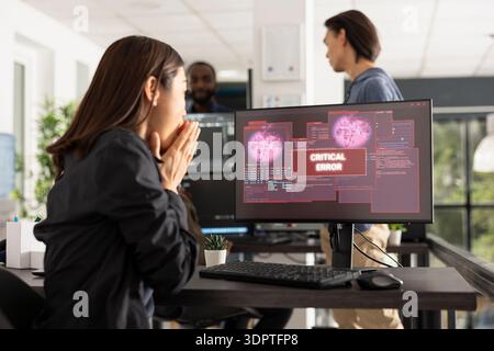 Portrait of female programmer reacting to alarming error alert on desktop monitor. Woman covers mouth in panic, representing software bugs, cyberattack threats and digital security crisis. Stock Photo