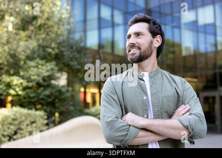 Smiling man standing with crossed arms outdoors, looking away happily with a modern office building and green trees in the background, showing a confident and successful attitude Stock Photo