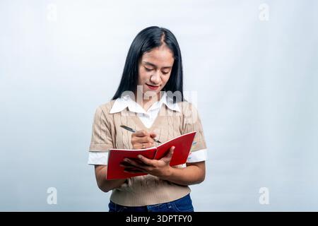 A young Asian woman in a beige vest and white shirt standing and writing notes in a red spiral notebook with a pen, isolated on a white background. Stock Photo