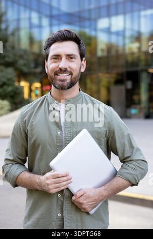 Smiling man standing confidently outdoors in front of a modern glass office building, holding a grey laptop symbolizing business, freelance work, and digital entrepreneurship in an urban context Stock Photo