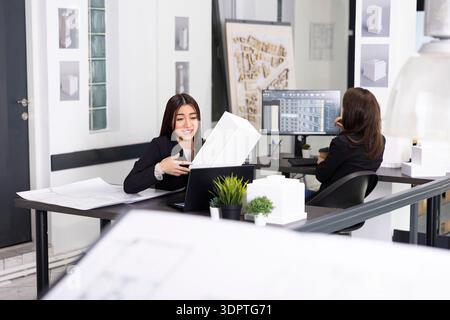 Smiling female architect holds small building model, reviewing project with contractors over video call. Asian designer connects through laptop to work with remote client to confirm architectural plans Stock Photo