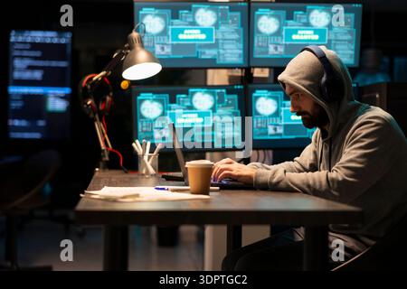 Male developer in hoodie works late at night with music, typing on laptop while monitoring multiple cybersecurity screens. Focused programmer with headphones, managing code and digital access data. Stock Photo