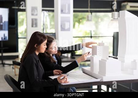 Focused female architects collaborate while analyzing white architectural model in modern office. Young professionals review project details, discussing structure, planning and design accuracy. Stock Photo