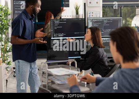 Group of developers sits around computers, discussing source code and system information during work meeting. Focused teamwork supports web development and problem solving in startup workspace. Stock Photo