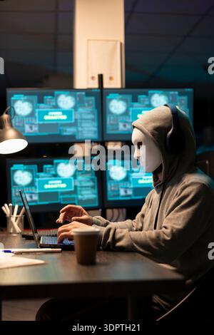 Anonymous person wearing hooded sweatshirt and headset types on laptop in office filled with access screens. Masked man creates malware code, attempting to breach security network in modern office. Stock Photo