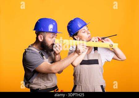Bearded man examines yellow measuring level as woman works with screw, showing teamwork in construction industry. Male builder focuses carefully on accuracy, precision and professional craftsmanship. Stock Photo
