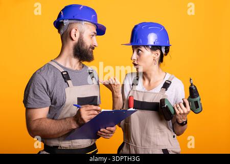 Caucasian maintenance crew collaborate on planning building repairs workflow. Male supervisor taking notes on clipboard as female contractor with drill provides updates on construction project. Stock Photo