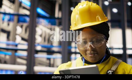 Upbeat professional working in smart factory oversees robot production lines, ensuring quality control. Industry 4.0 plant expert monitoring automated conveyor systems using notebook, camera B Stock Photo