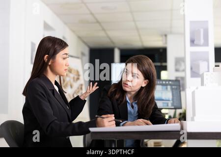 Diverse female professionals work together in office, analyzing floor plans and coordinating tasks for upcoming construction project. Women architects discussing building designs at desk. Stock Photo