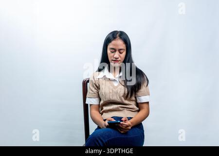 A young Asian woman in a beige vest sitting on a chair, looking down at her mobile phone with a serious and focused expression on a white background. Stock Photo