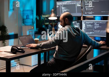 Bearded male programmer stretches across desk to adjust both laptop and desktop monitors. Focused software developer works late in modern tech workspace filled with screens and coding interfaces. Stock Photo