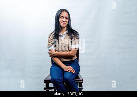 A happy young Asian woman with long black hair sitting on a chair, looking over her shoulder with a bright smile on a white studio background. Stock Photo