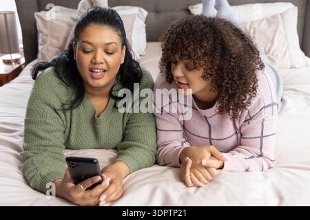 Diverse female friends lying on stomachs on bed in green and pink grid sweaters, sharing smartphone Stock Photo
