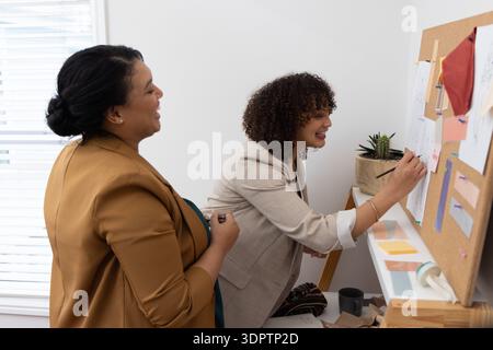 Diverse female team in blazers collaborating at studio corkboard, marking fabric swatches and notes Stock Photo