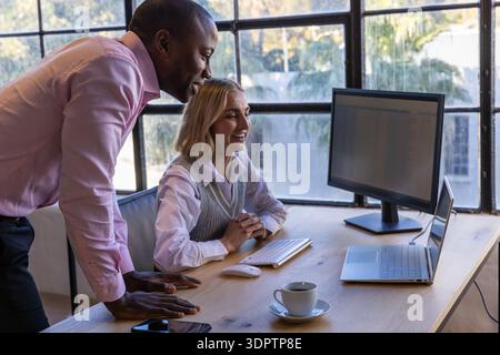 Diverse coworkers leaning over desk, interacting with laptop, coffee cup at office Stock Photo