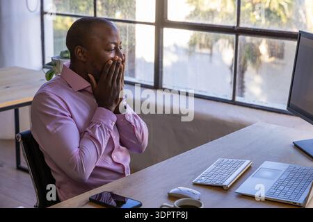 African american man covering mouth and viewing monitor at office desk with coffee mug, copy space Stock Photo