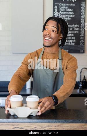 African American male barista handing two lidded coffee cups in cardboard carrier at cafe counter Stock Photo